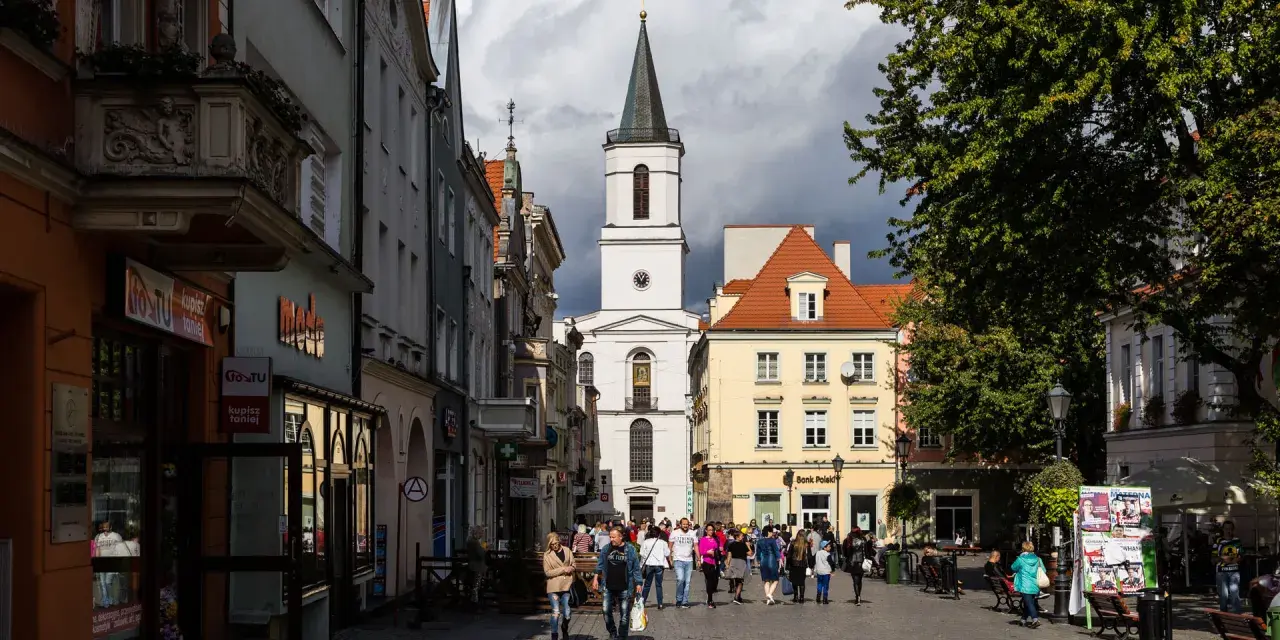 Ulica w jednym z miast na zachodzie Polski, z kościołem w tle i ludźmi spacerującymi po rynku.