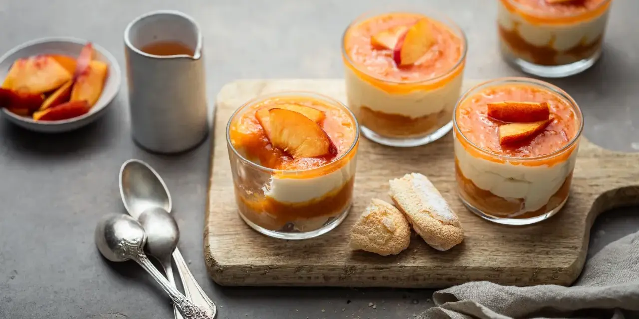 Delizioso tiramisù alle pesche in coppette, con pesche fresche e savoiardi su tagliere di legno.