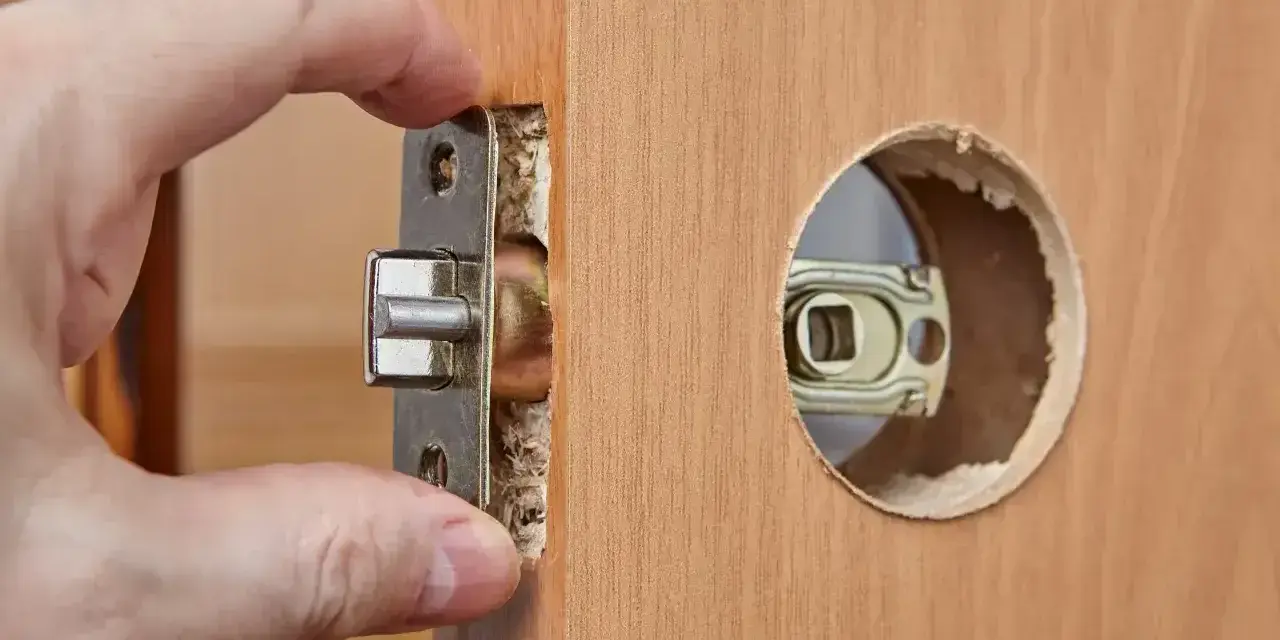 Dłoń montuje mechanizm zamka w drewnianych drzwiach. Dowiedz się, ile kosztuje wymiana zamka w drzwiach.