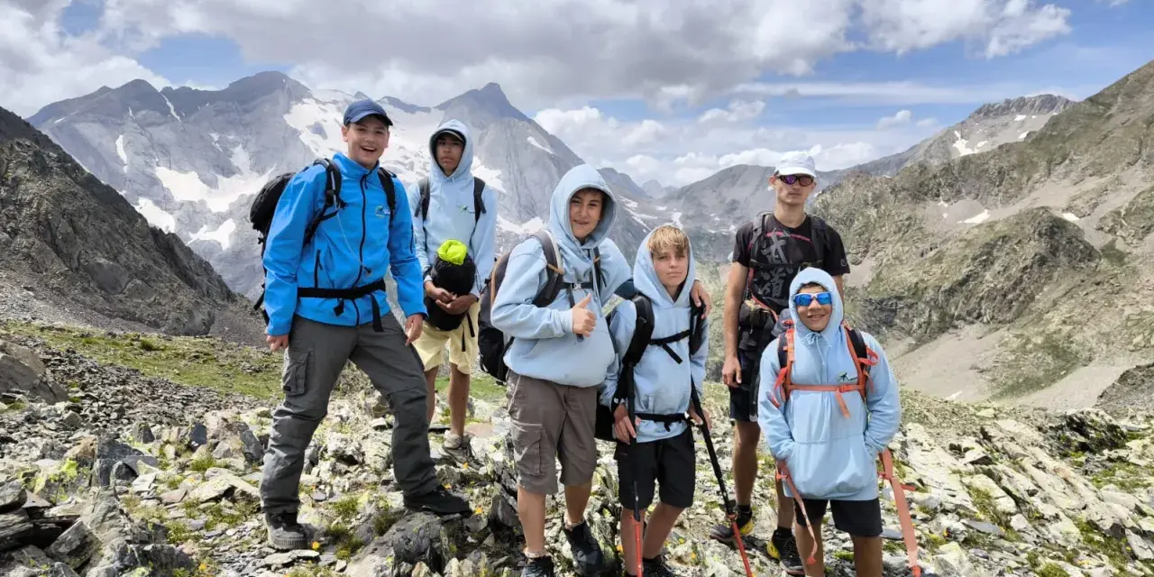 Groupe d'enfants et d'un adulte en randonnée en montagne, souriants, avec des sacs à dos et des bâtons de marche.