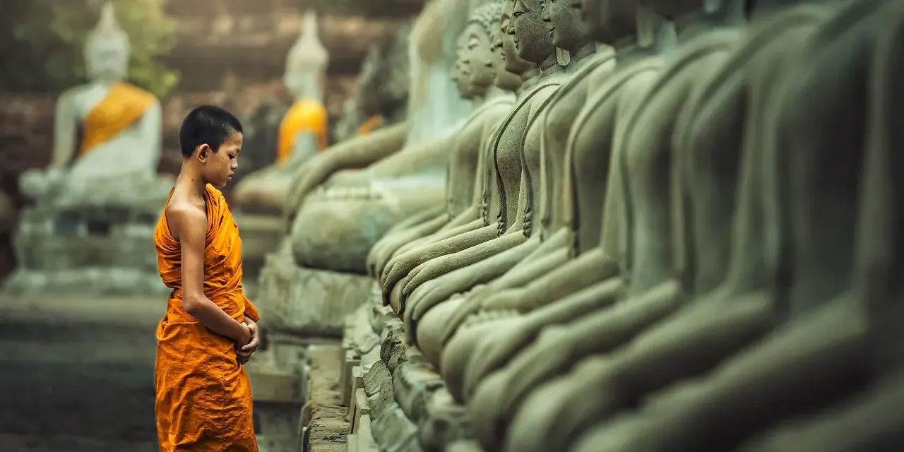 A young monk in orange robes stands before a row of Buddha statues, a scene evoking the ancient roots of what is the oldest religion in the world.