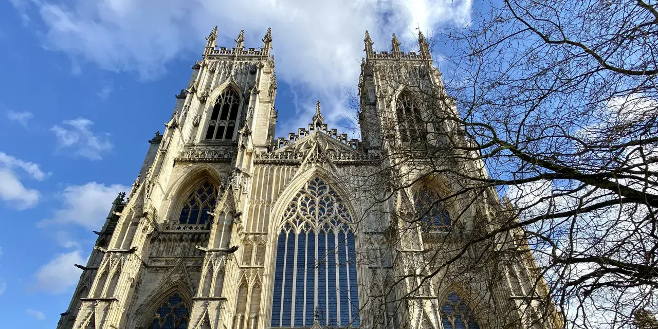 The majestic York Minster, a Gothic cathedral, stands tall against a bright blue sky with scattered clouds. Bare tree branches frame the intricate stonework and towering spires.