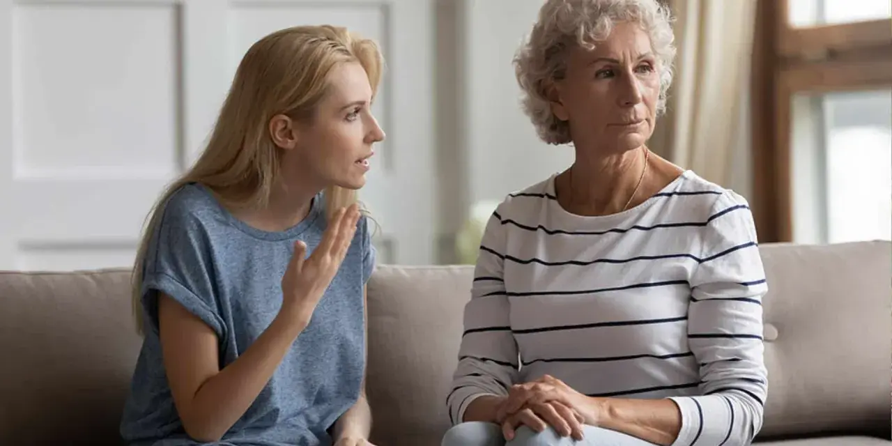 Junge Frau spricht vorwurfsvoll zu ihrer Mutter, die abgewandt sitzt. Die Mutter beleidigt die erwachsene Tochter.