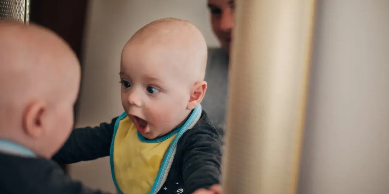 Ein Baby mit gelbem Lätzchen schaut überrascht in den Spiegel. Es ist fasziniert von seinem Spiegelbild, ein Moment, wann sagen Kinder "Ich".