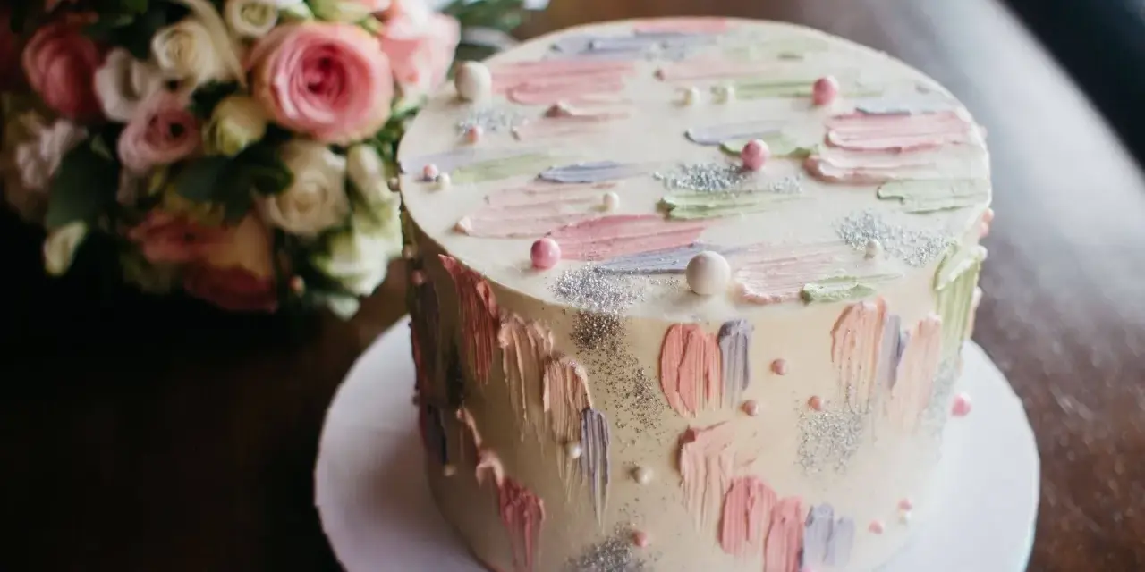 Oto tort na 20 osób, ważący około 2 kg, ozdobiony pastelowymi pociągnięciami pędzla i perełkami. Obok bukiet różowych kwiatów.