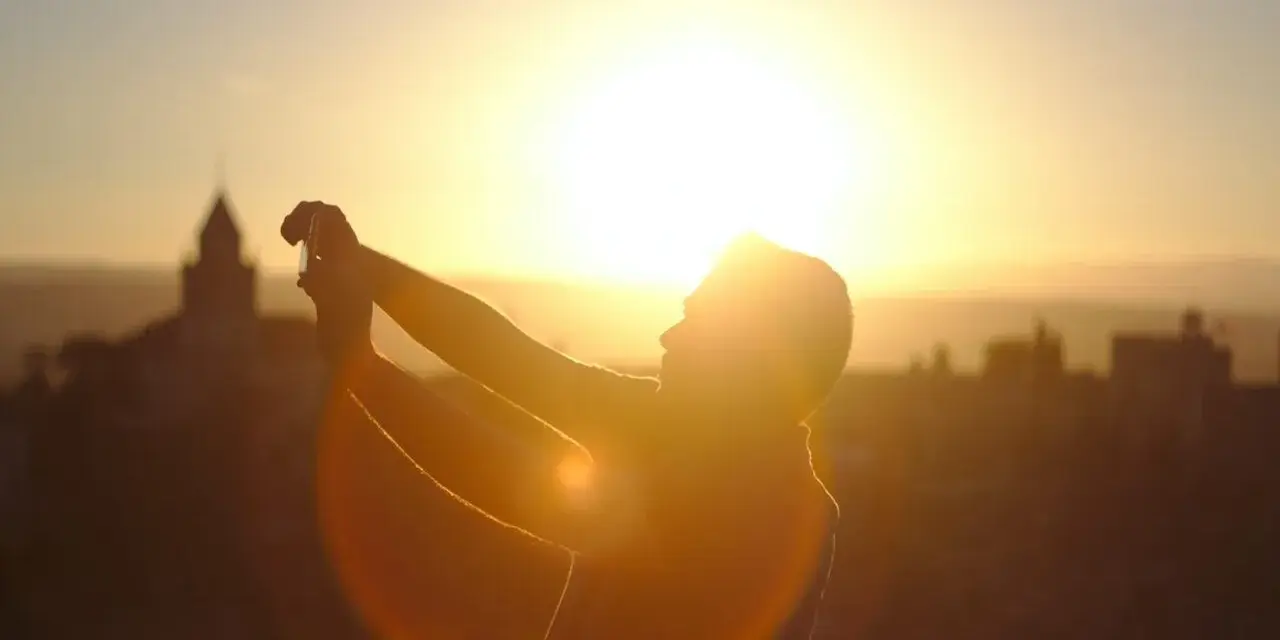 Silhouette d'une personne prenant une photo contre jour avec son téléphone, le soleil éclatant à l'horizon derrière des bâtiments.