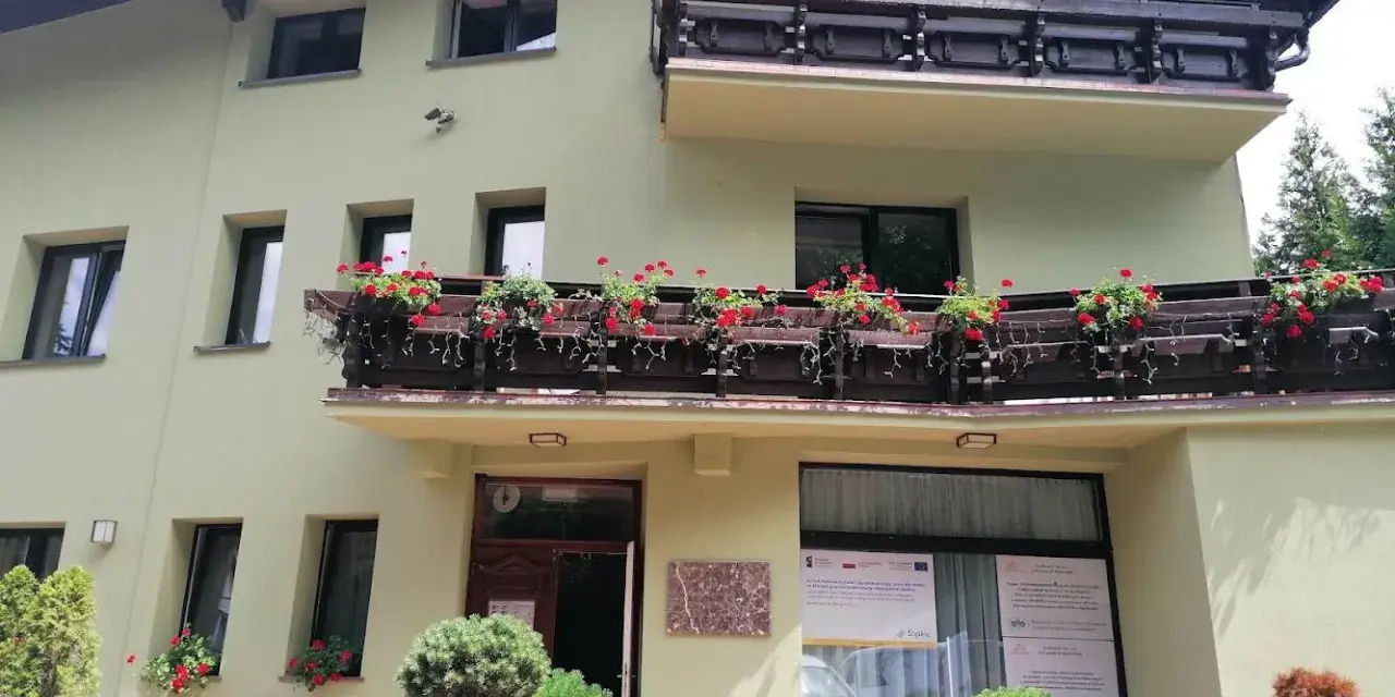 Jasnozielony hotel na błoniach z balkonem pełnym czerwonych kwiatów.