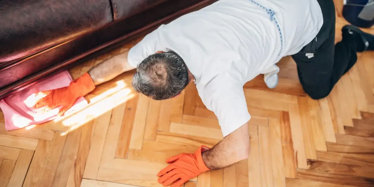 Man in orange gloves cleaning wooden floor with a pink cloth.