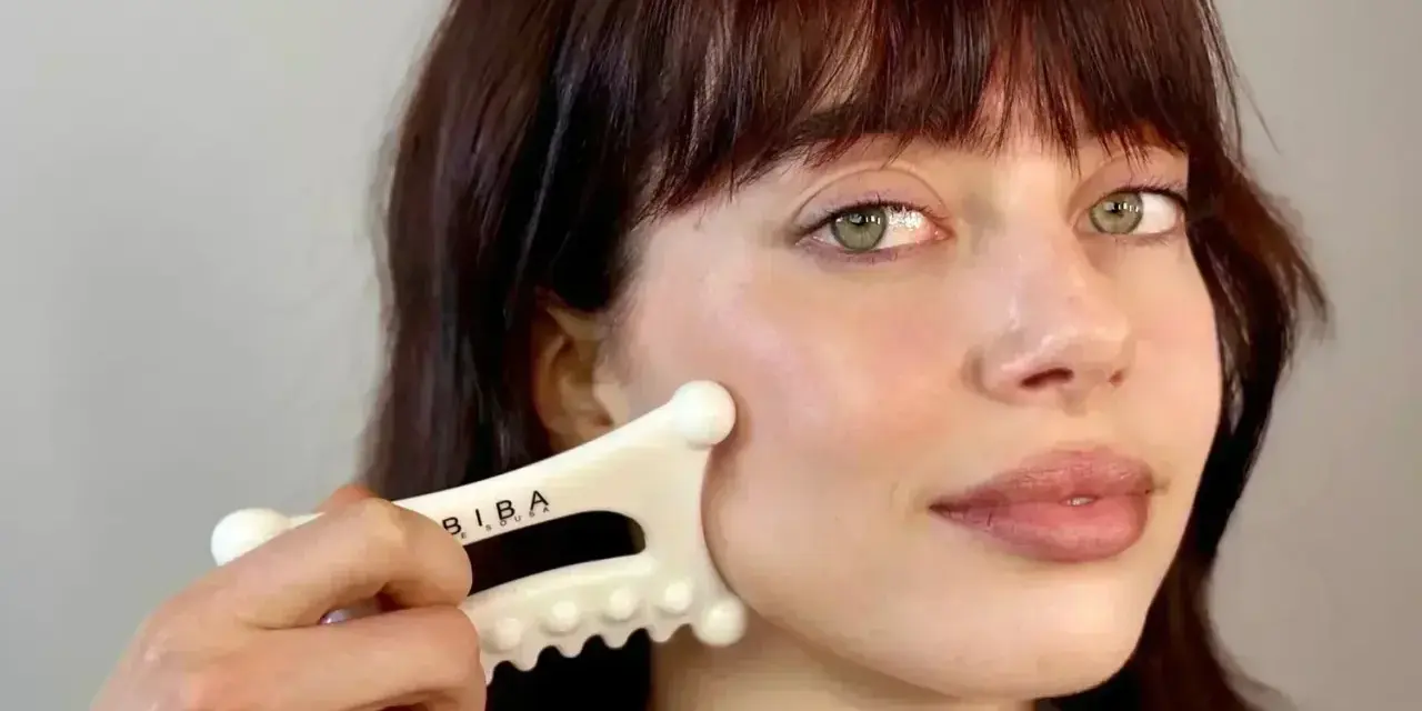 A woman with bangs and green eyes performs a facial fascia massage using a white gua sha tool.
