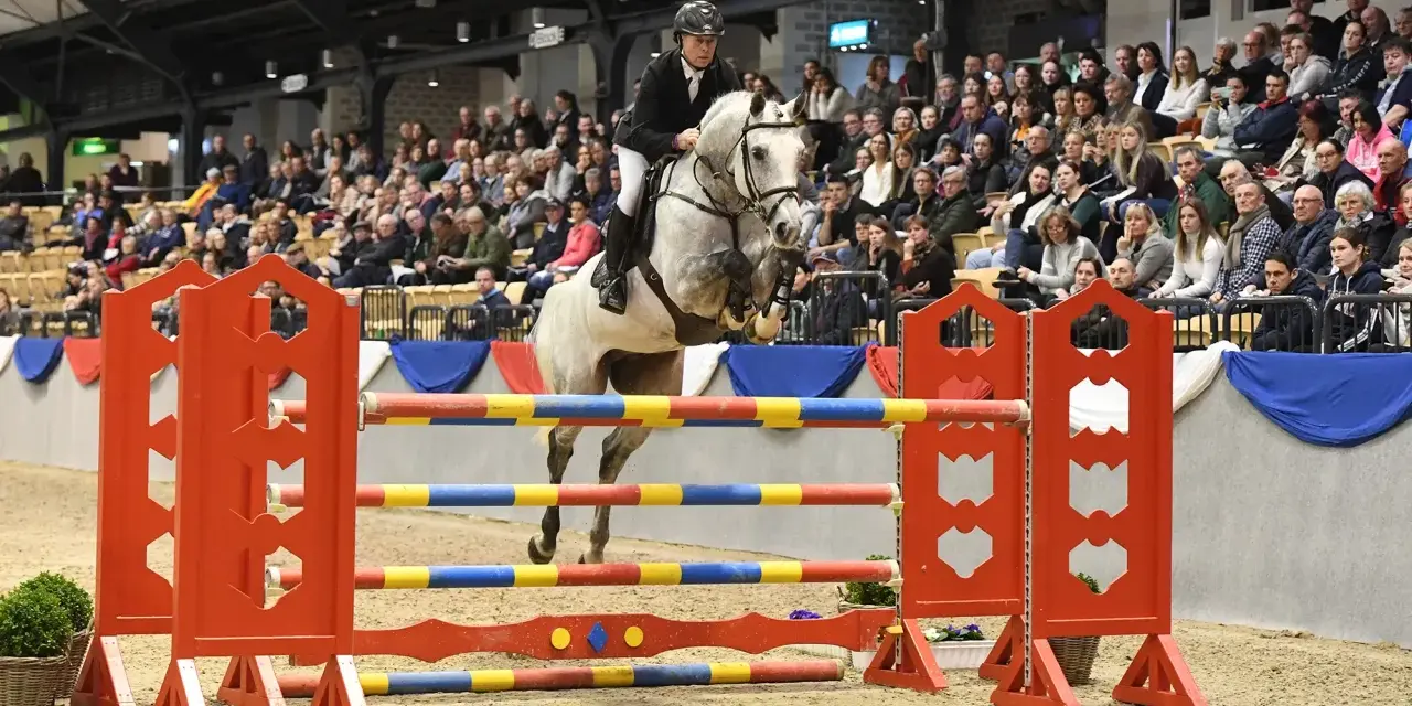 Un cheval blanc, un véritable con air etalon, saute une barrière colorée sous les regards d'une foule attentive.