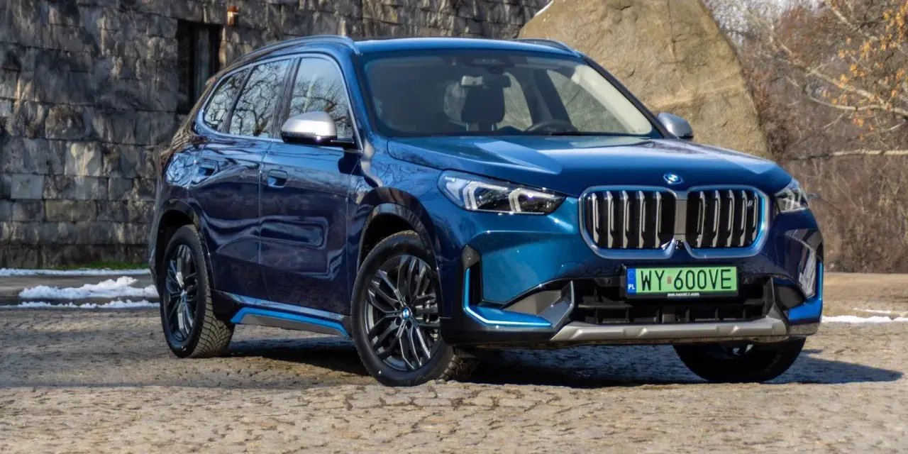 Niebieskie BMW iX1 stoi na kamienistej nawierzchni przed kamienną ścianą.