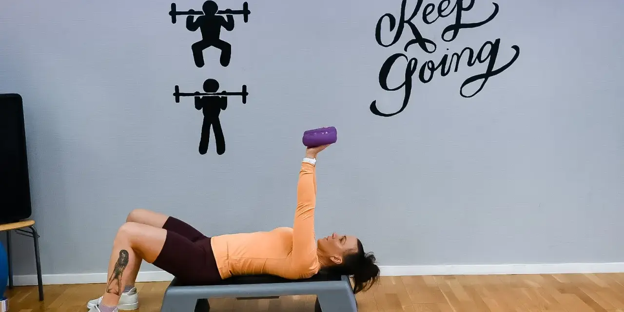 Kvinna tränar med en lila hantel på en stepbräda. Väggen har texten "Keep Going" och silhuetter av styrkelyft.