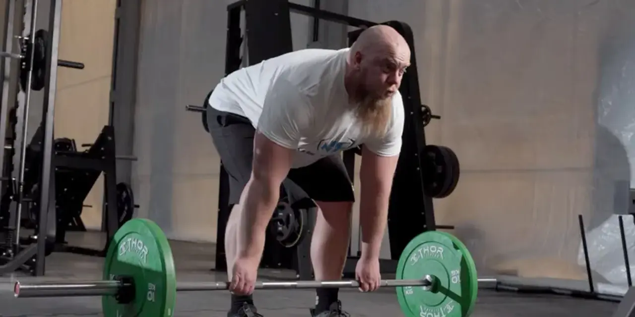 Man med skägg utför raka marklyft med hantlar i ett gym.