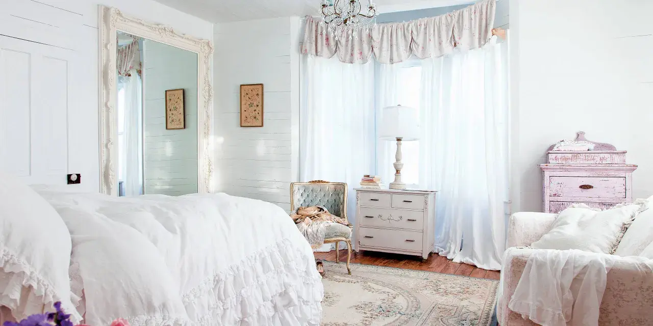 Dormitorio con cama blanca y volantes, espejo grande, cómoda antigua y sillón. Estilo shabby chic.