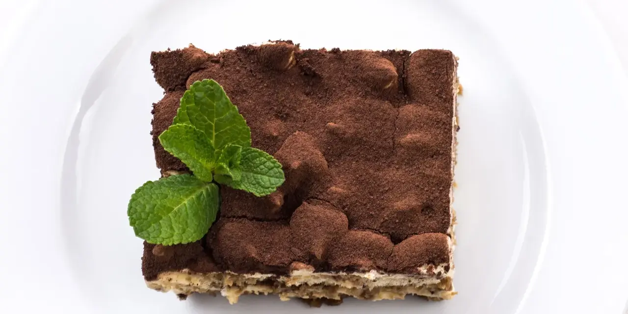 Pyszne tiramisu bez alkoholu, posypane kakao i udekorowane listkami mięty, na białym talerzu.