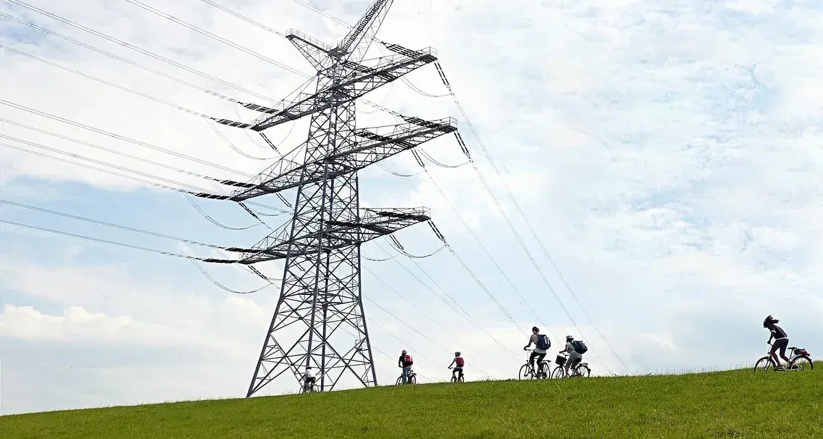 Grupa rowerzystów przemierza zielone wzgórze pod potężną konstrukcją linii 110 kV.