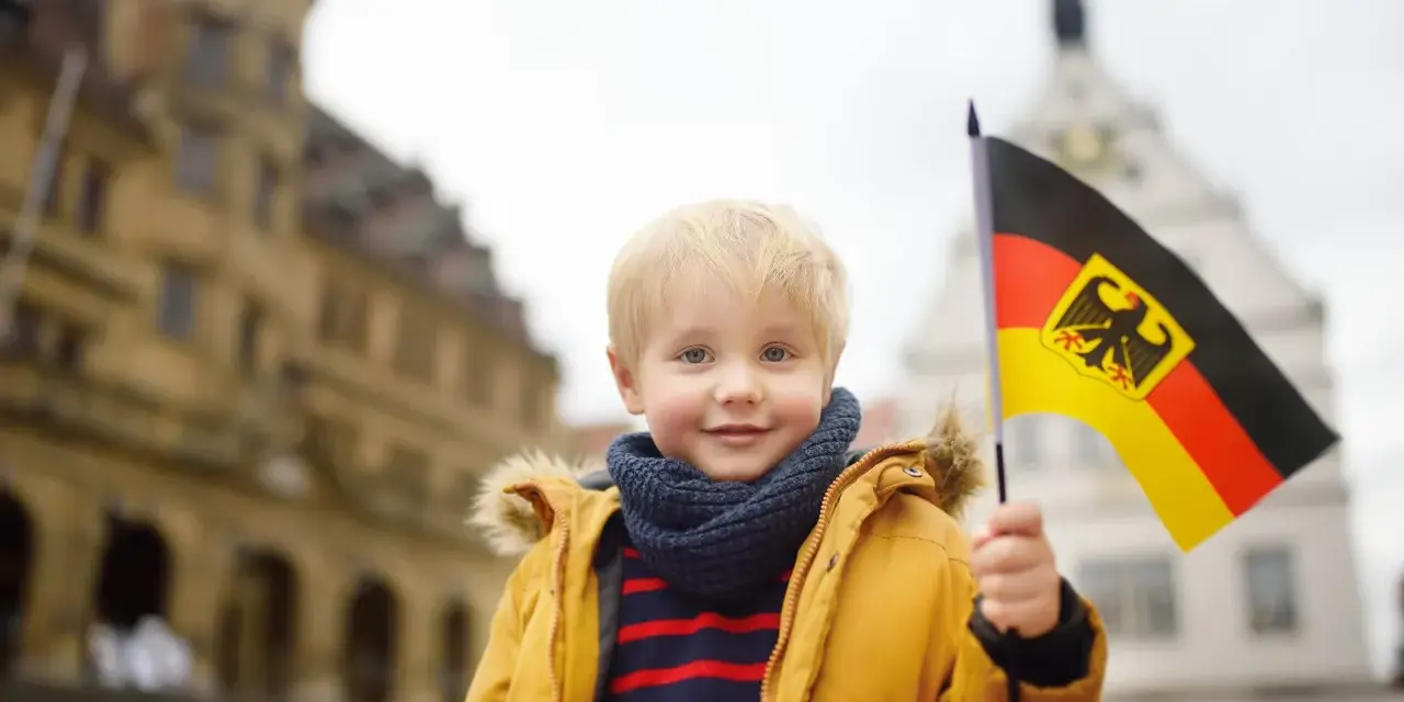 Uśmiechnięty chłopiec z blond włosami trzyma flagę Niemiec. W tle widać zabytkowe budynki, przypominające te, które można spotkać w niemieckich miasteczkach, gdzie znajdują się szkoły.