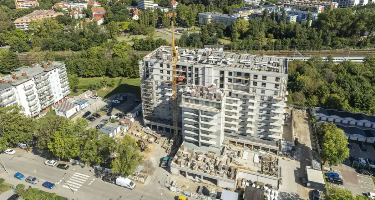 Budowa bloku mieszkalnego w Kołobrzegu. Widok z lotu ptaka na plac budowy, dźwig, samochody i okoliczne budynki. Ceny mieszkań Kołobrzeg mogą być różne.