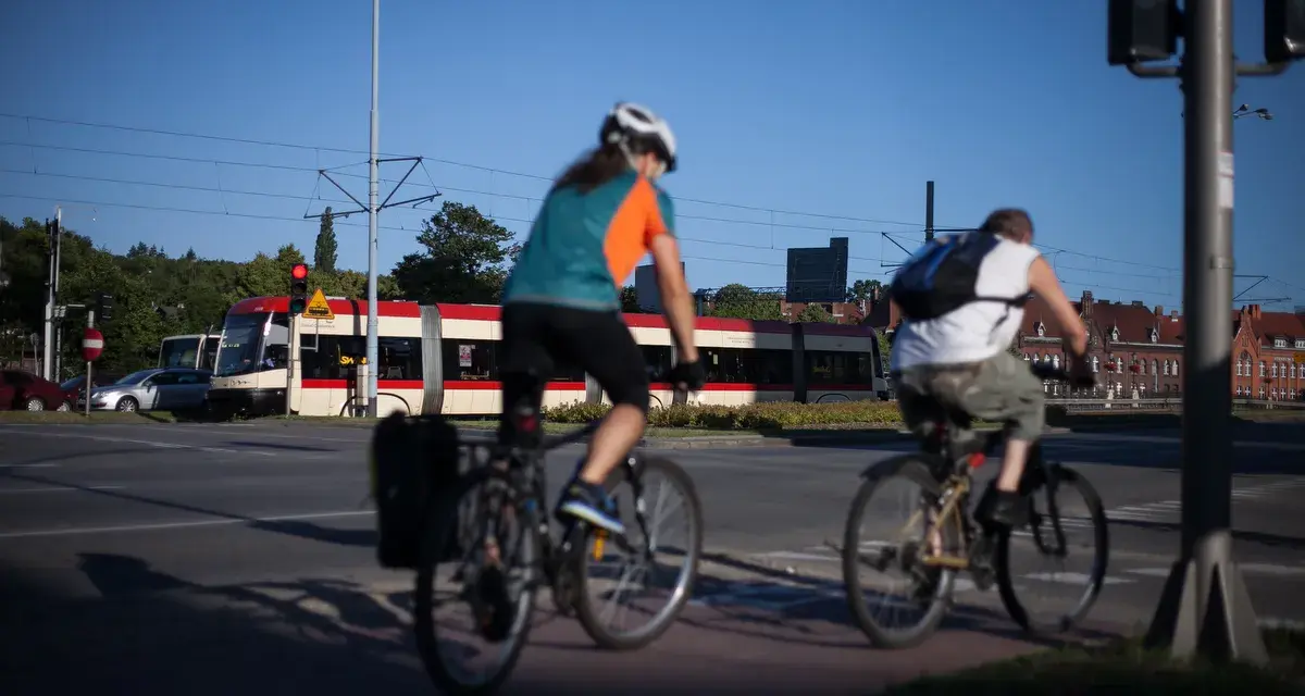 Rowerzyści jadący wzdłuż torów tramwajowych, w tle czerwono-biały tramwaj. Trasa rowerowa wiodąca w kierunku Westerplatte.