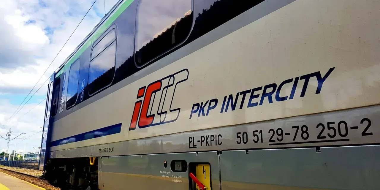 Pociąg PKP Intercity z napisem "Warszawa Poznań" na tle błękitnego nieba z chmurami.