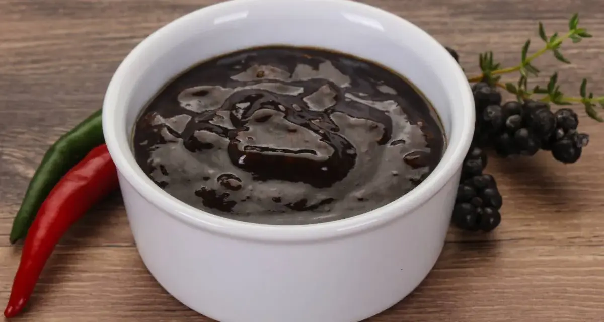 Gęsty, ciemny sos pieprzowy w białej miseczce, obok czerwona i zielona papryczka chili oraz gałązka jeżyn.