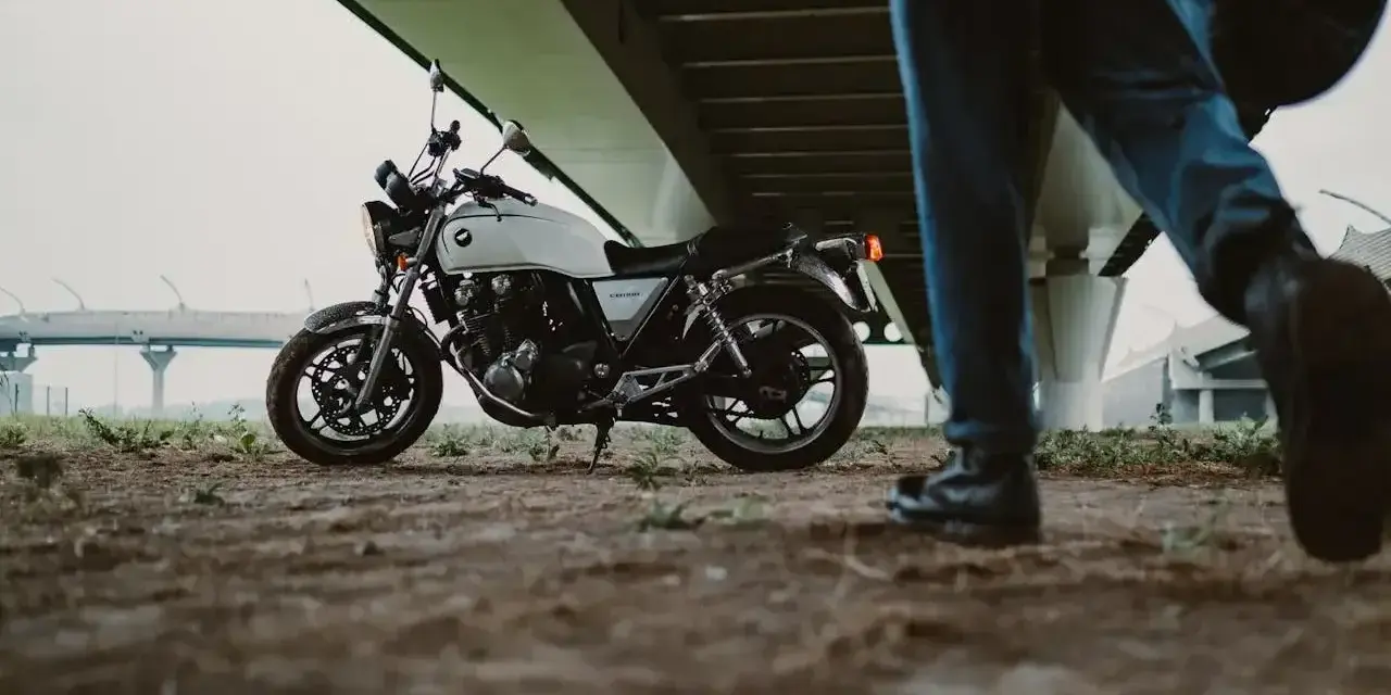 Biały motocykl klasyczny, kategoria A2, pod mostem. Obok przechodzi motocyklista z kaskiem.