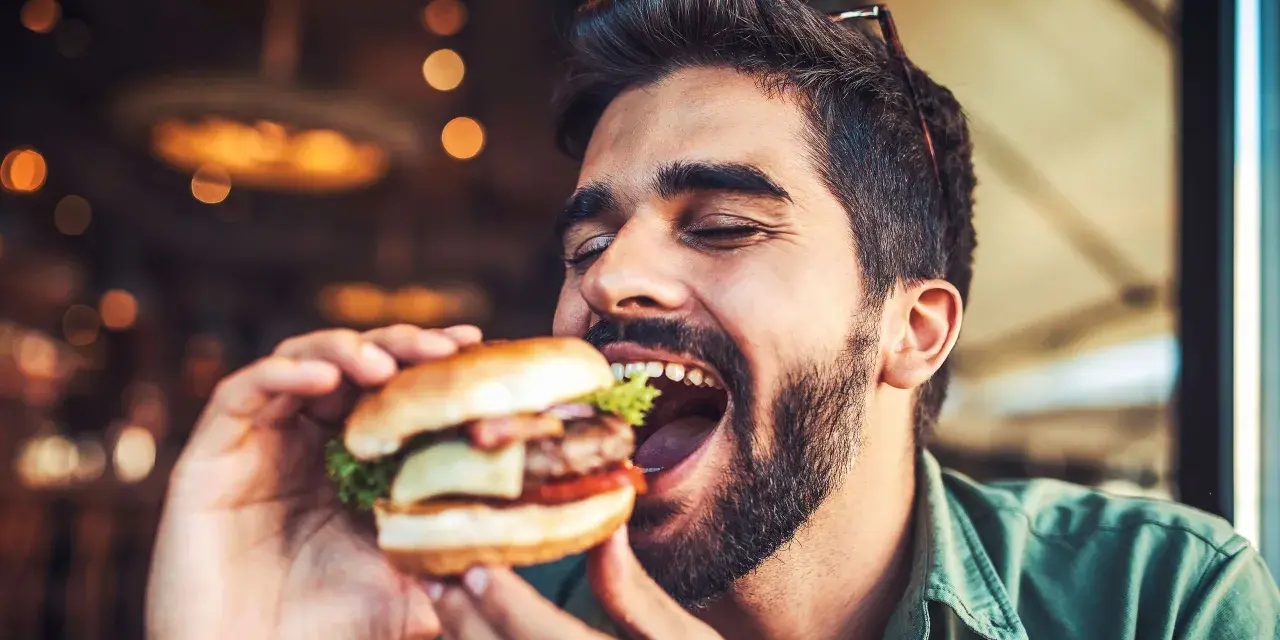 Hombre disfrutando un jugoso **cheat meal** de hamburguesa, con los ojos cerrados por el placer.