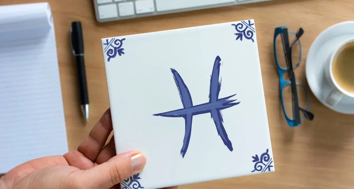 Dłoń trzyma kwadratową płytkę z symbolem ryby, znakiem zodiaku. Na biurku leżą notatnik, długopis, okulary i kawa.