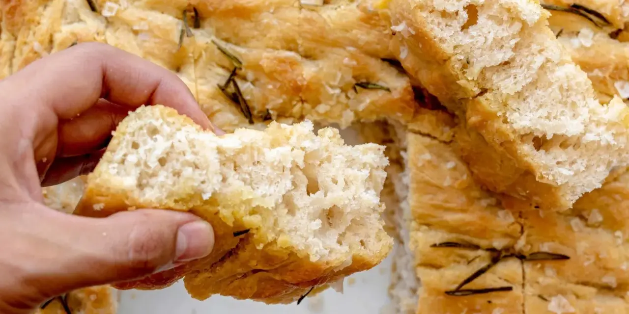 Mano rompiendo un trozo de focaccia casera con romero y sal gruesa. ¡Una delicia de esta focaccia receta!