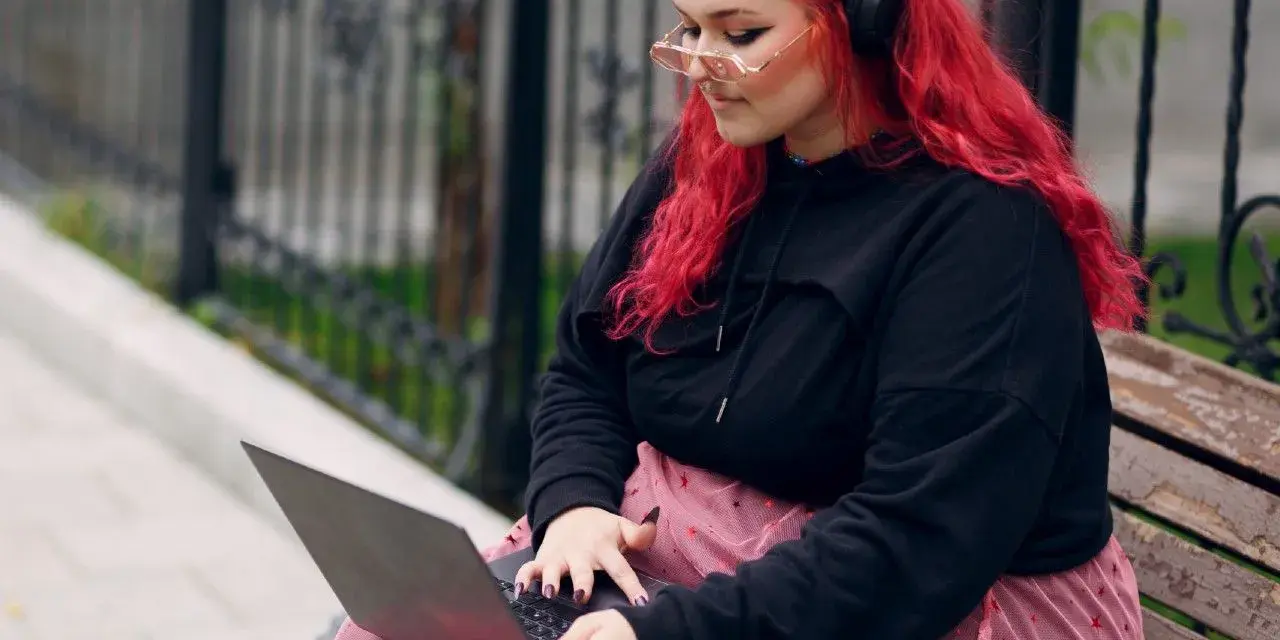 Młoda babista z ognistymi włosami i słuchawkami na uszach pracuje na laptopie, siedząc na ławce w parku.