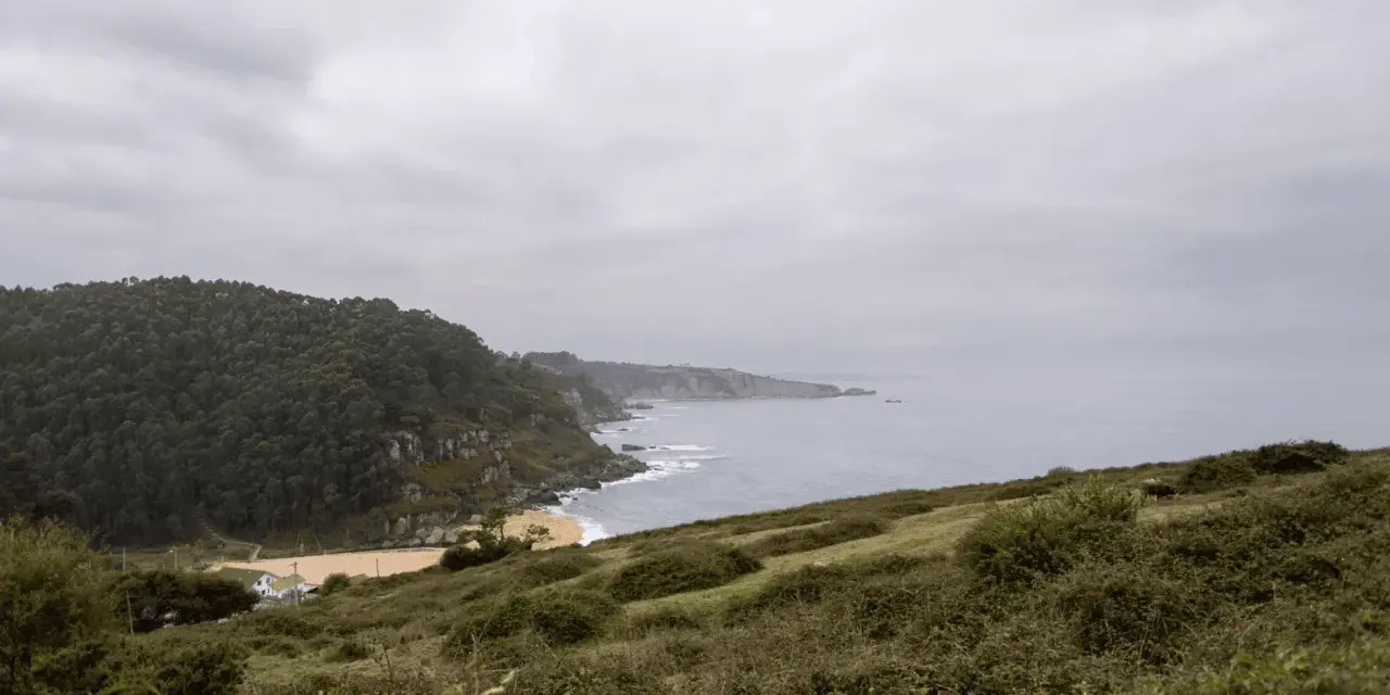 Paisaje costero con acantilados verdes y mar gris. Ideal para senderismo en Villaviciosa, con una playa dorada y una casa blanca.