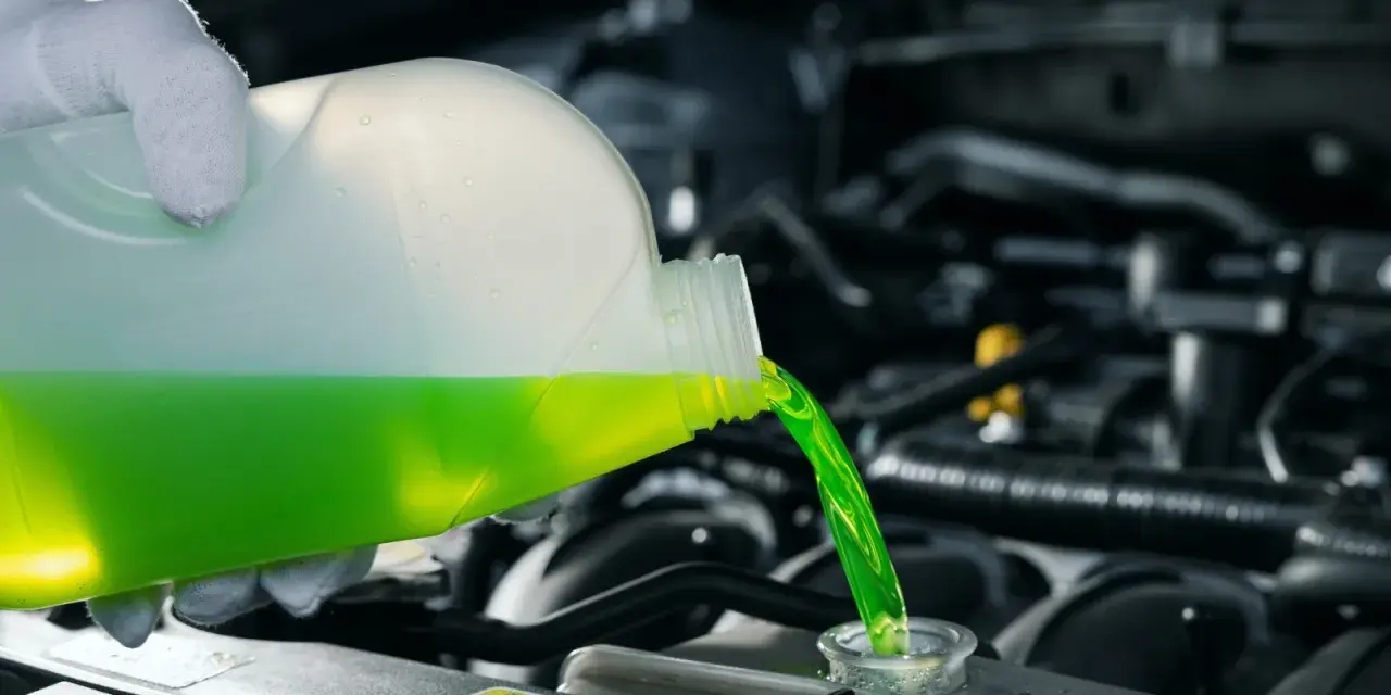 A gloved hand pours green antifreeze and coolant into a car's engine.
