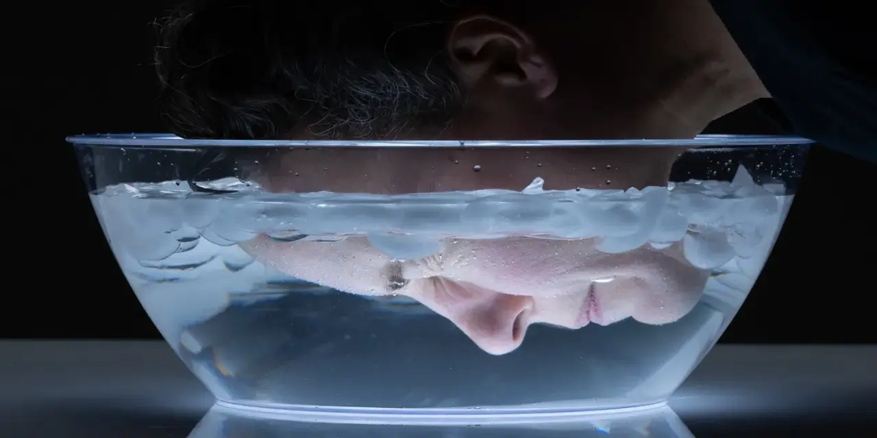 A person submerges their face in a clear bowl filled with water and ice cubes for a facial ice bath.