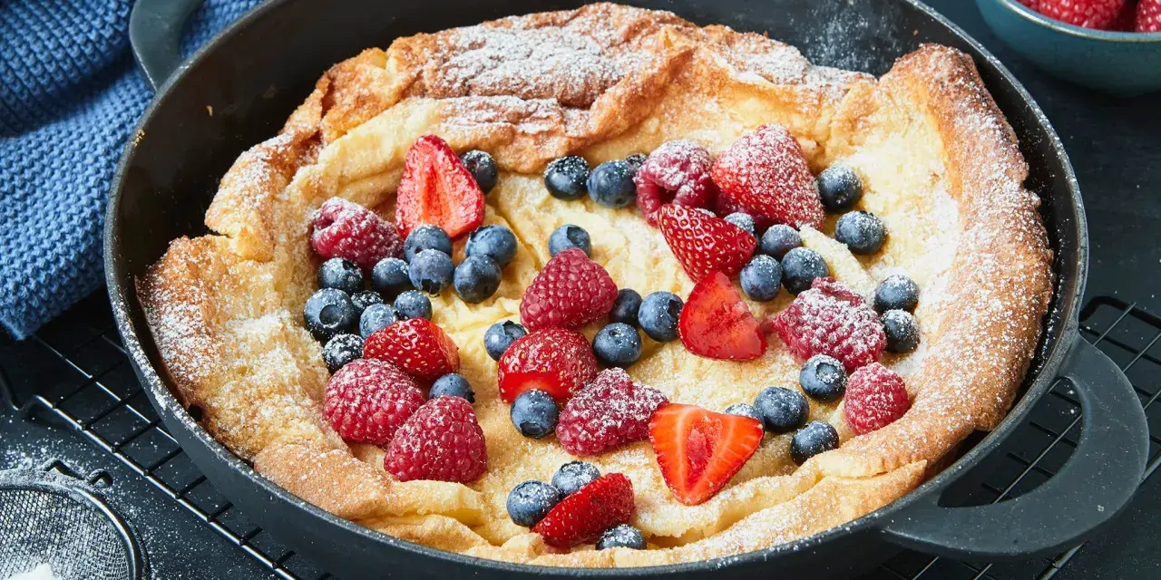Ein goldbrauner Dutch Baby mit Puderzucker und frischen Beeren, serviert in einer gusseisernen Pfanne.