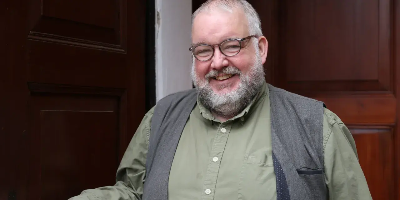 Ein freundlicher evangelischer Pfarrer mit Brille und Bart lächelt in die Kamera, vor einer dunklen Holztür stehend.