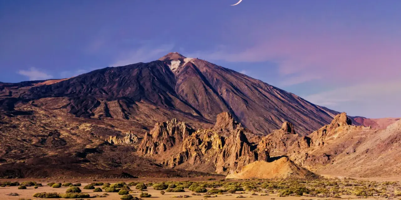 Krajobraz w parku narodowym Teide z księżycem w kształcie sierpa na fioletowym niebie. Wulkan góruje nad skalistym terenem.