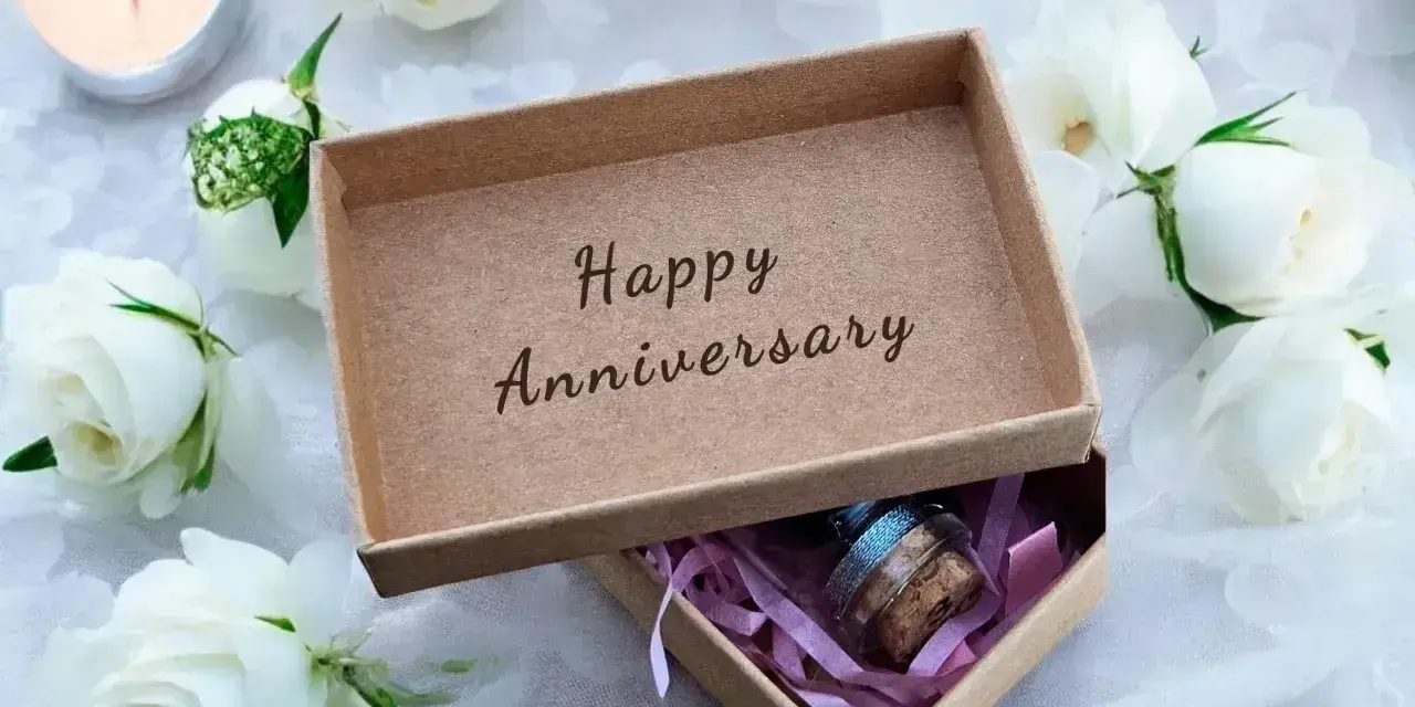 Otwarta szkatułka z napisem "Happy Anniversary" i małym prezentem w środku, otoczona białymi różami i płonącą świecą. Idealna na rocznice ślubu.