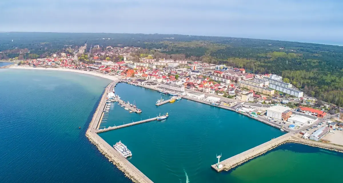 Widok z lotu ptaka na malowniczy hel port z licznymi łodziami i statkami. W tle rozciąga się miasto i las.