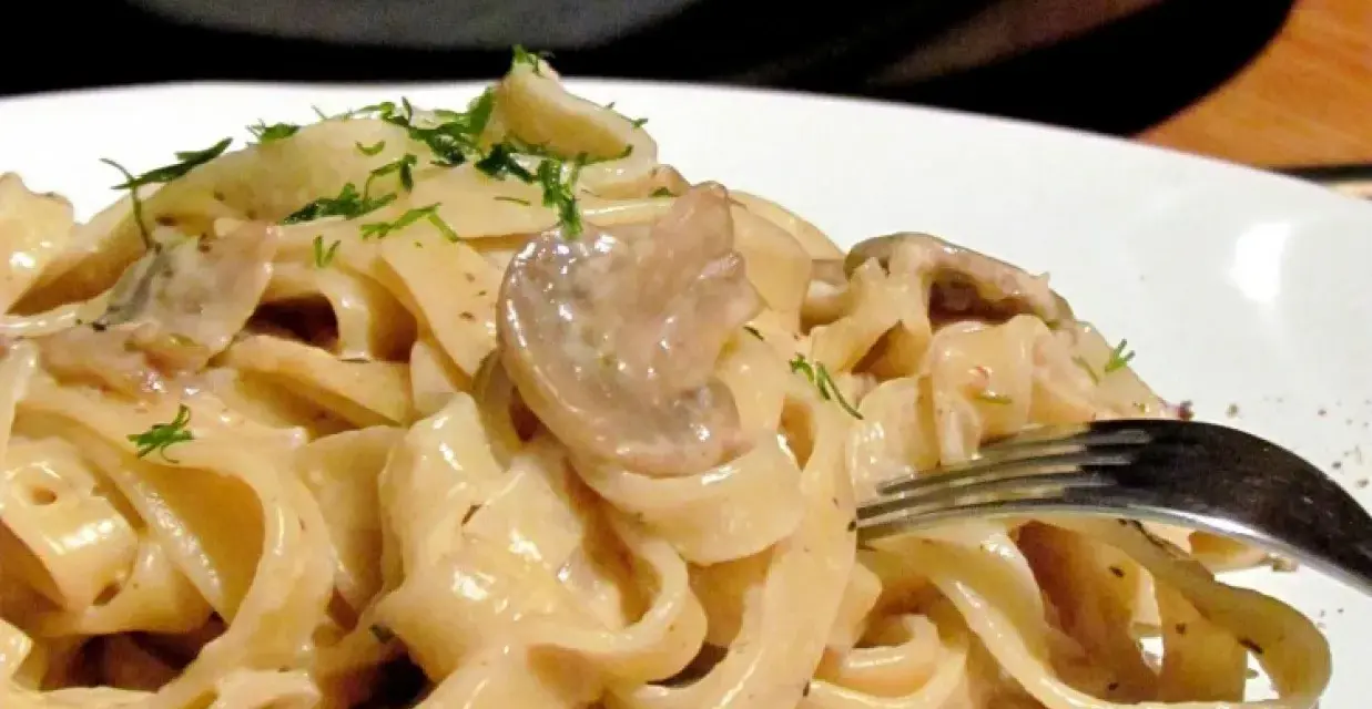Tagiatelle z grzybami w kremowym sosie, posypane świeżym koperkiem. Widok z bliska na talerz pełen pysznego makaronu.