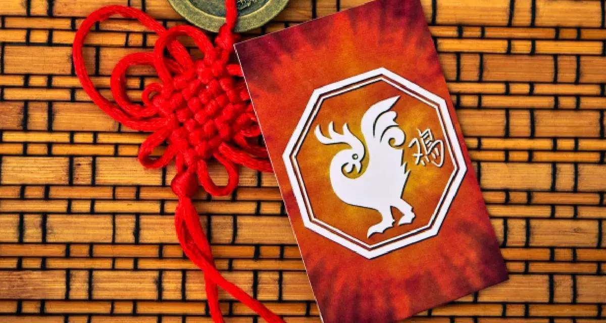 Czerwony chiński węzeł i karta z białym kogutem, symbolem 1993 chińskiego znaku zodiaku, na tle bambusowym.