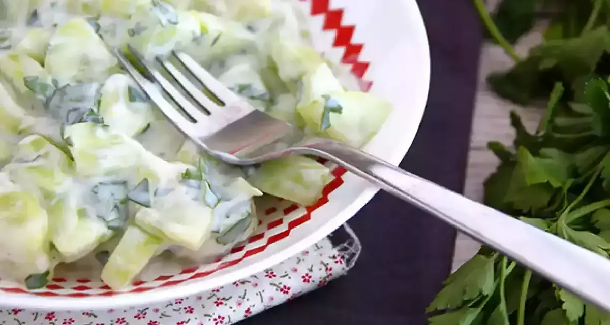Ensalada cremosa de pepino y yogur con hierbas frescas en un plato blanco con borde rojo.