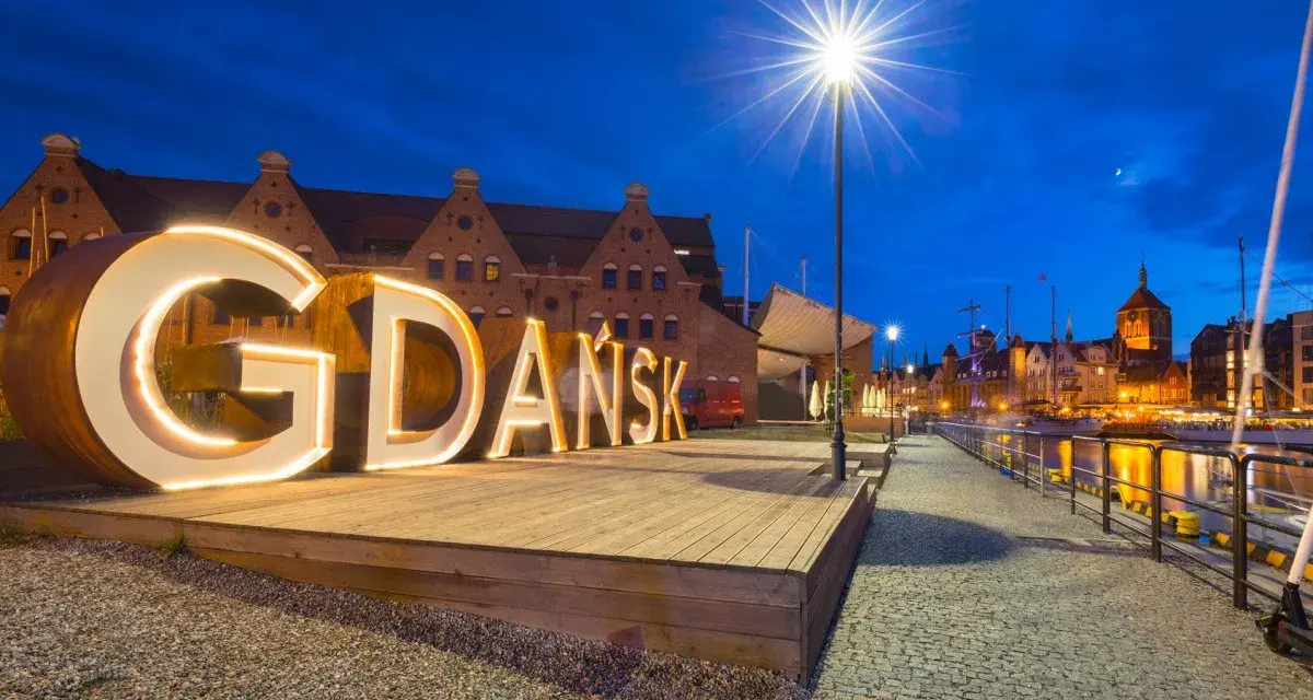 Podświetlany napis "GDAŃSK" na tle nocnej panoramy miasta, zapraszający do odkrywania atrakcji Trójmiasta.
