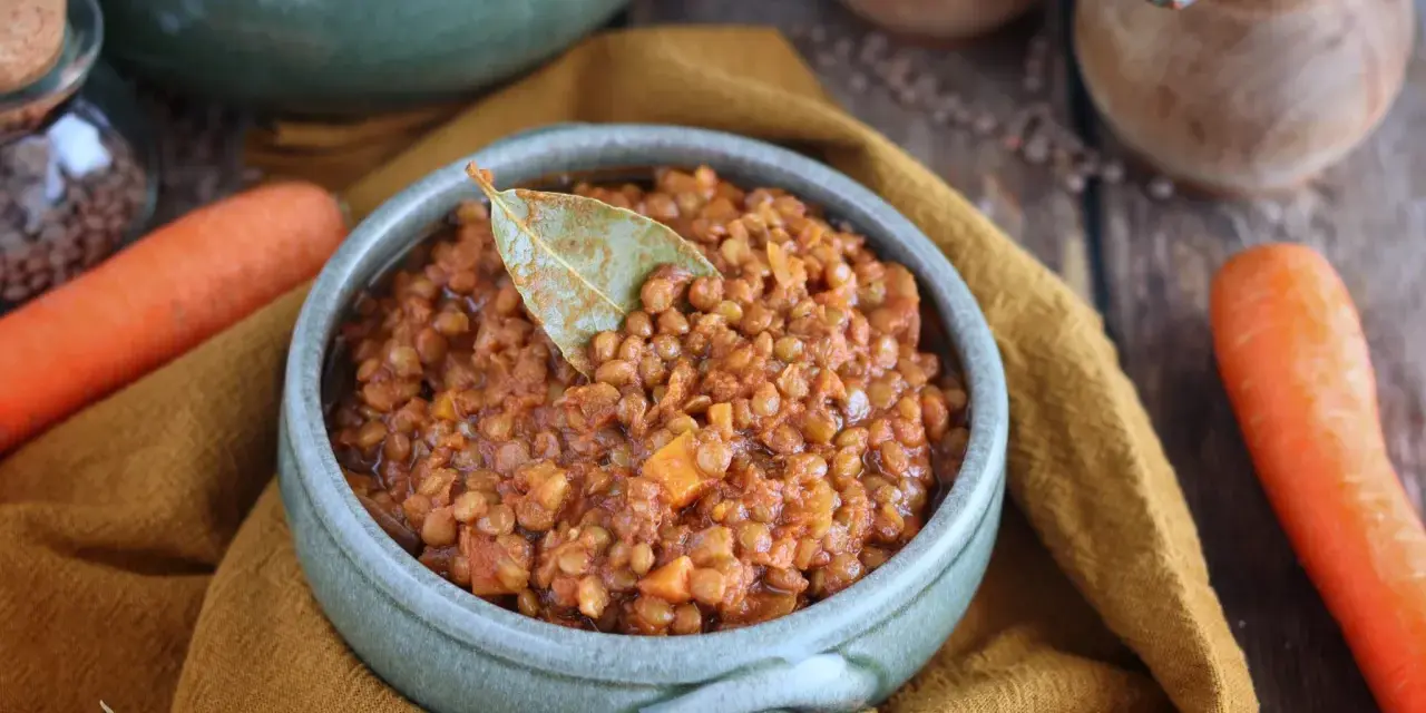 Un invitante ragù di lenticchie, profumato con alloro, pronto per essere gustato. Accanto, carote fresche e cipolla.