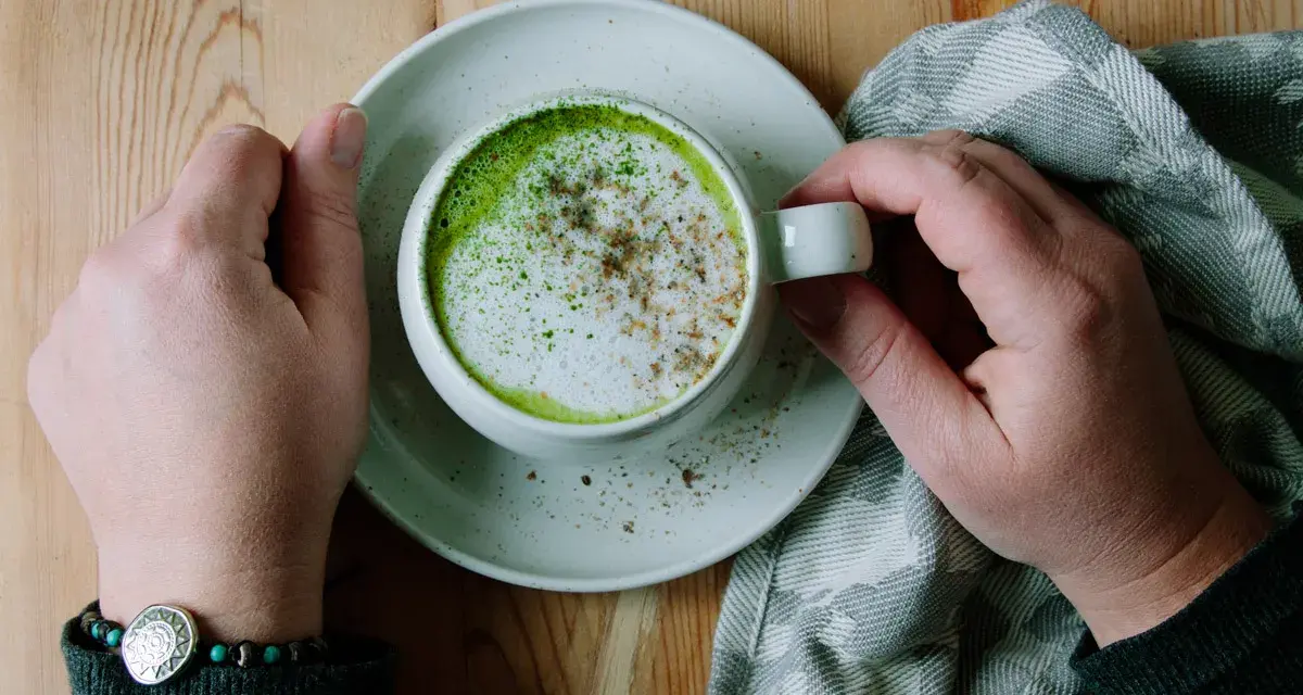 Händerna håller en kopp med en krämig matcha latte, toppad med skum och lite kryddor. En mysig stund med en grön dryck.