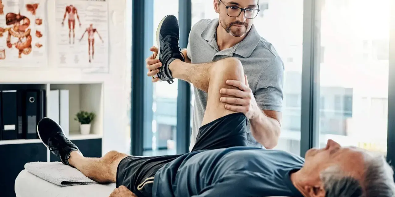 Ein Physiotherapeut dehnt das Bein eines Patienten. Der Patient liegt auf einer Liege, während der Therapeut sein Knie beugt. Wann Physiotherapie? Hier wird geholfen.