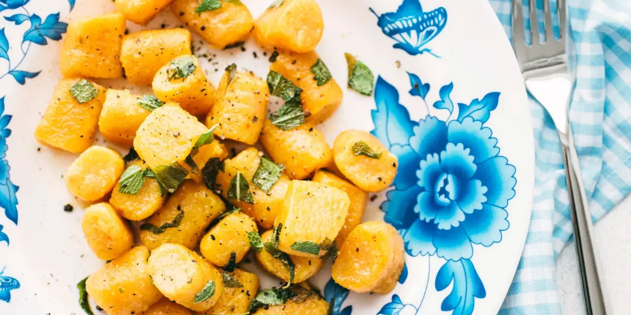 Gnocchi di zucca dorati, conditi con erbe aromatiche e pepe nero, su un piatto decorato con fiori blu.