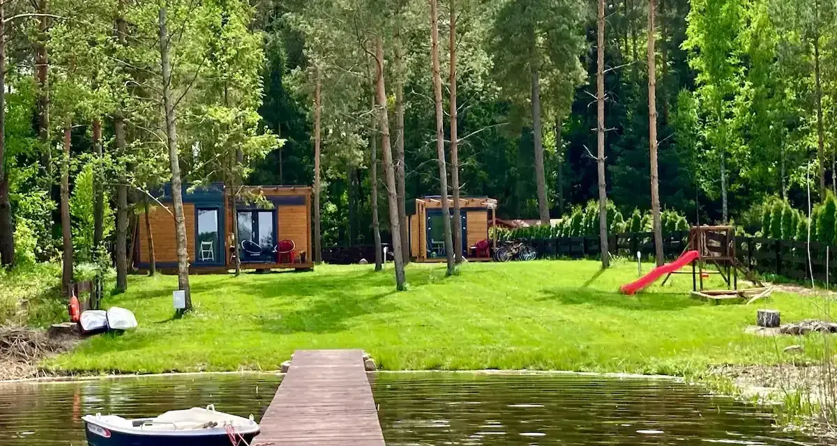 Drewniany pomost nad jeziorem Bardze, z łódką przycumowaną do brzegu. W tle domki letniskowe i plac zabaw.