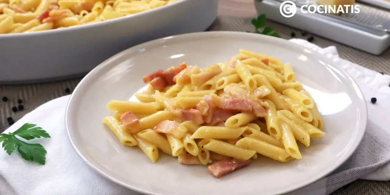 Plato de macarrones a la carbonara cremosos con trozos de panceta. En el fondo, un bol con más pasta y un rallador de queso.