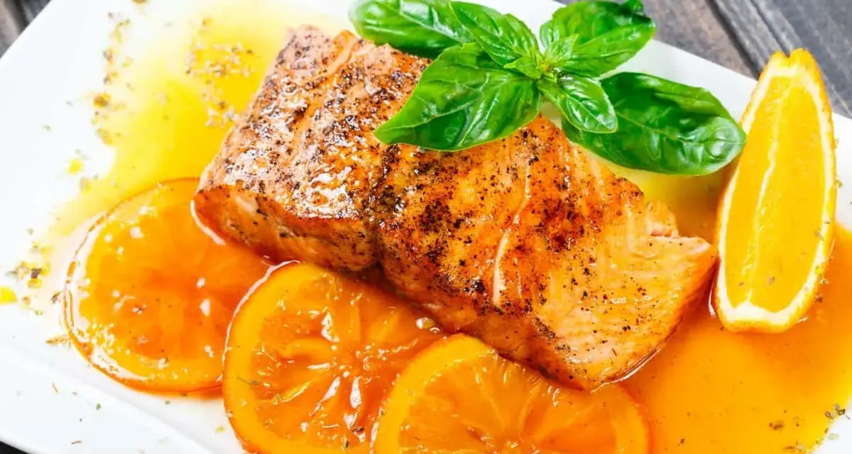 Delicioso salmón a la naranja, servido con rodajas de naranja fresca y hojas de albahaca.