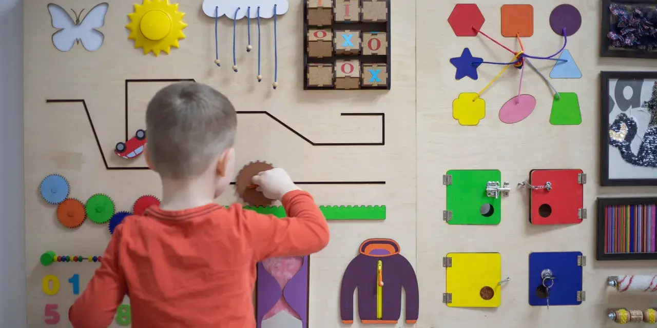 Chłopiec bawi się na tablicy edukacyjnej, rozwijając umiejętności manualne zgodnie z metodą Montessori. Widoczne są kolorowe kształty, zębatki i zamek.